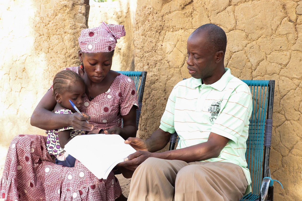 A female member of our project in Mali sat holding her baby with a Tree Aid partner staff member, filling in a form